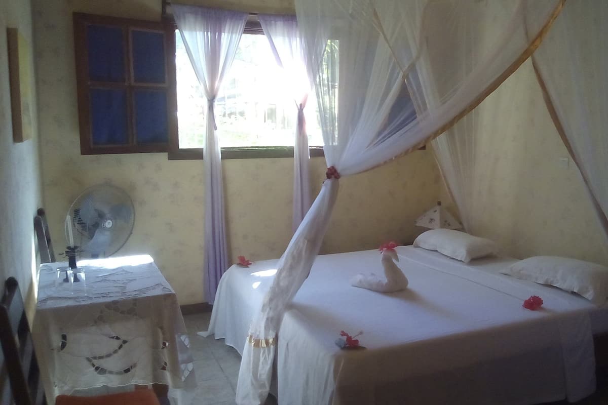 Una habitación con cama, una mesa, un ventilador y una ventana con cortinas.