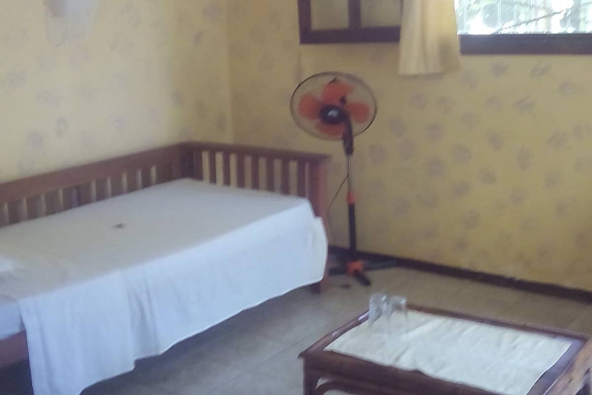 Una habitación con cama, una mesa, un ventilador y una ventana con cortinas.