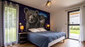 Une chambre avec un grand lit, une table de chevet avec une lampe, une fenêtre avec des rideaux et une fresque représentant un ours sur le mur.