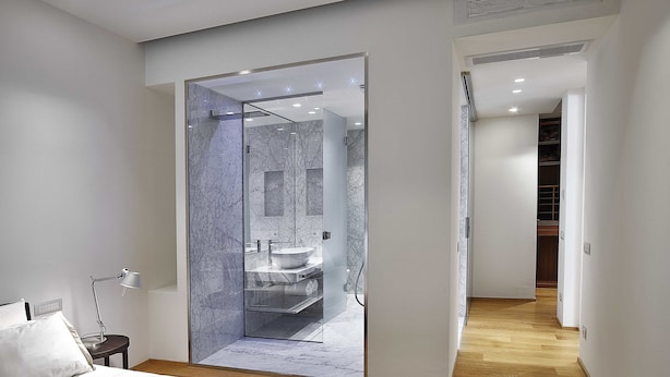 A modern bedroom with a large bed, a bedside table, and a nightstand. There is a glass-enclosed shower area with a marble wall and a sink. The room has wooden flooring and white walls. - Lario Apartment (Como)