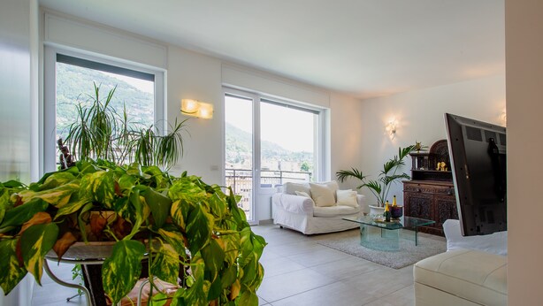 A modern living room with a white sofa, a glass coffee table, and a TV. There are large windows with a view of a mountain and a balcony. - Victoria Apartment (Como)