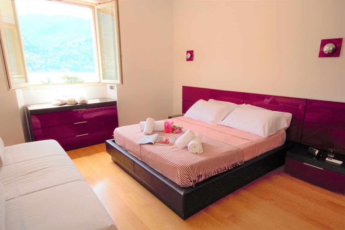 A neatly made bed with a pink headboard, a bedside table with a remote control, and a window with a view of a cityscape.