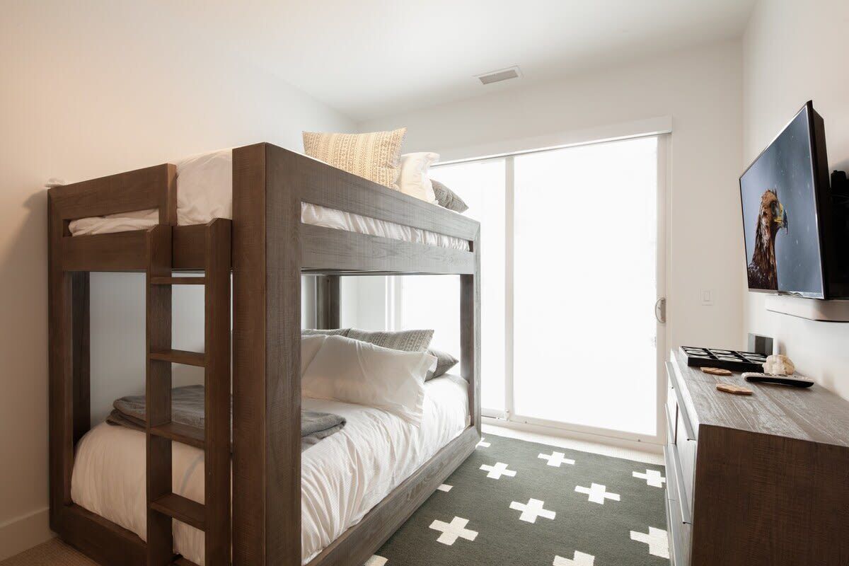 A bunk bed with a wooden frame and white bedding, a television on a wooden stand, and a cross-patterned rug on the floor.