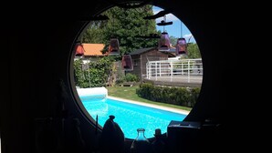 Vista a través de un ventanal circular que muestra una piscina, una terraza de madera y una casa con techo rojo.