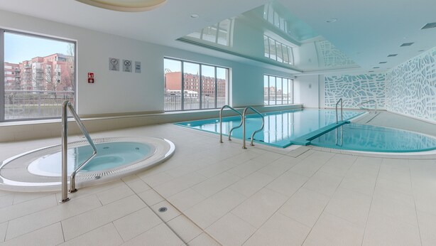 Une piscine intérieure moderne avec des parois en verre transparent, un jacuzzi et une vue sur les bâtiments environnants.