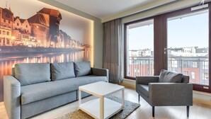 Un salon moderne avec un canapé gris, une table basse blanche et un fauteuil gris. Une grande fenêtre offre une vue sur des immeubles et une rivière.