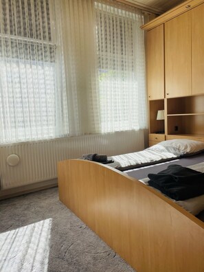 Une chambre à coucher avec un lit, un radiateur, une fenêtre avec des rideaux légers et une armoire en bois.
