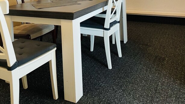 Een eethoek met een houten tafel en witte stoelen, een wit kastje met planken, en een decoratieve ankerhanger aan de muur.