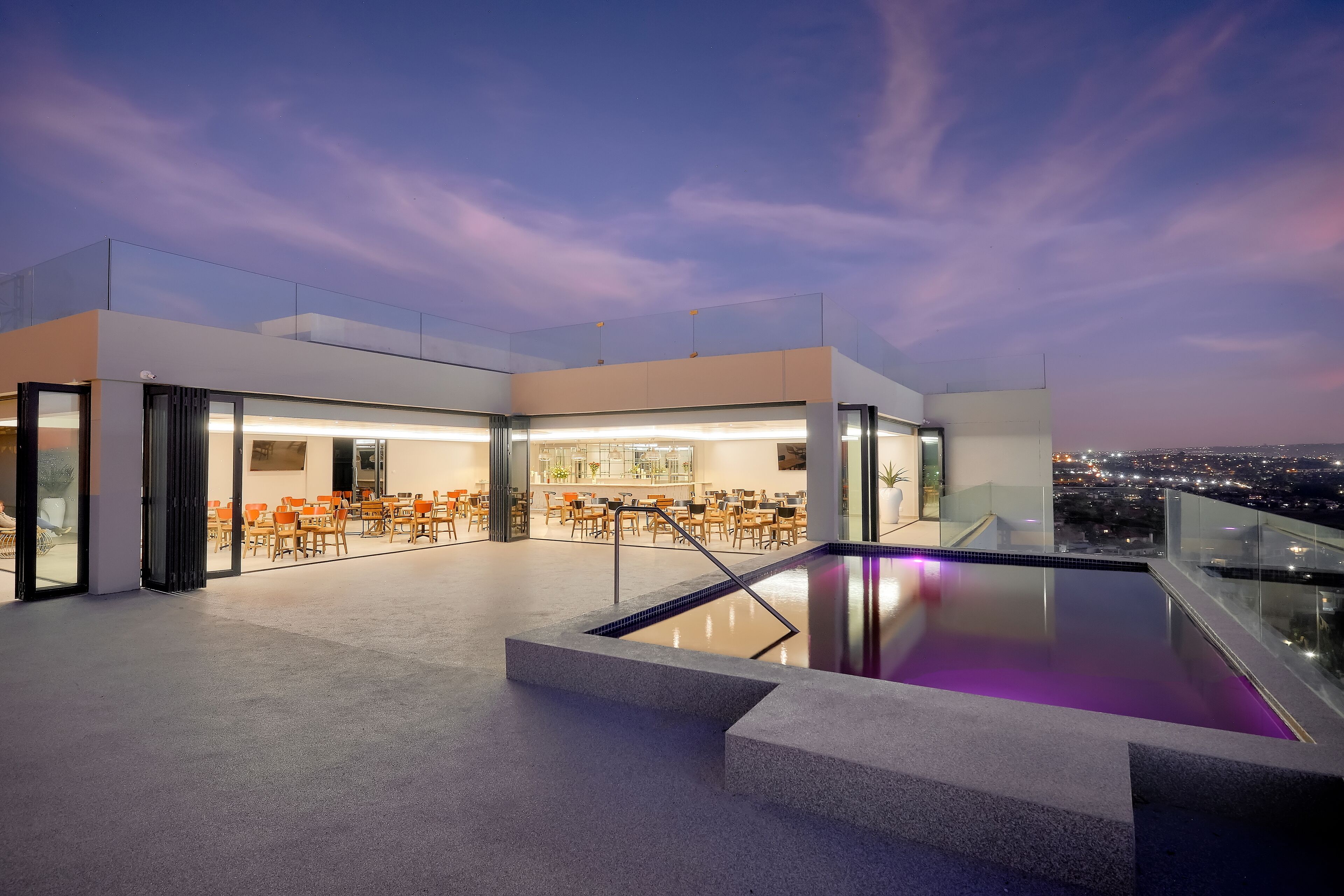 A rooftop terrace with a pool, seating area, and a view of the city at dusk.