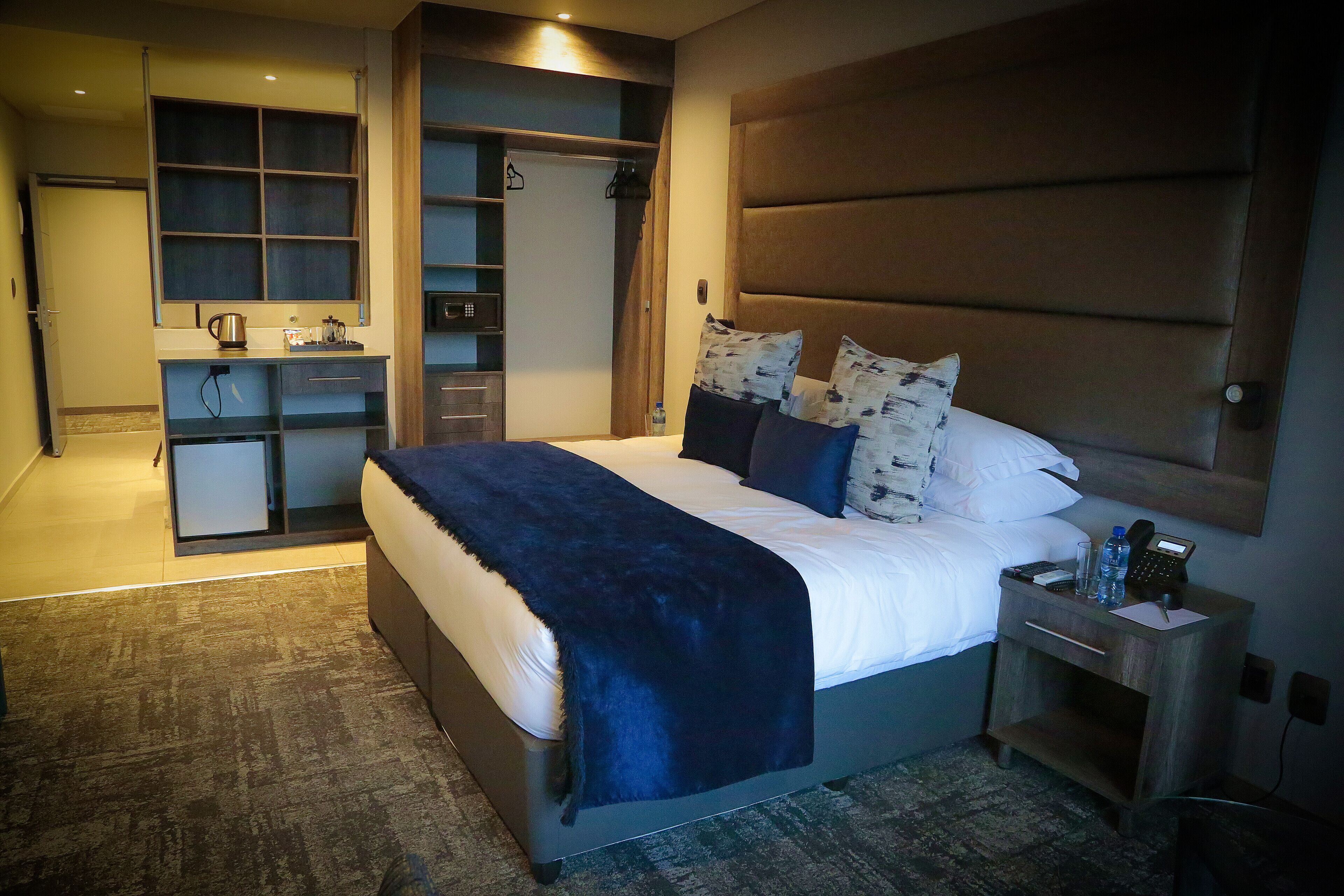A hotel room with a large bed, a bedside table with a phone and water bottle, a wardrobe with a mirror, and a small desk with a kettle.