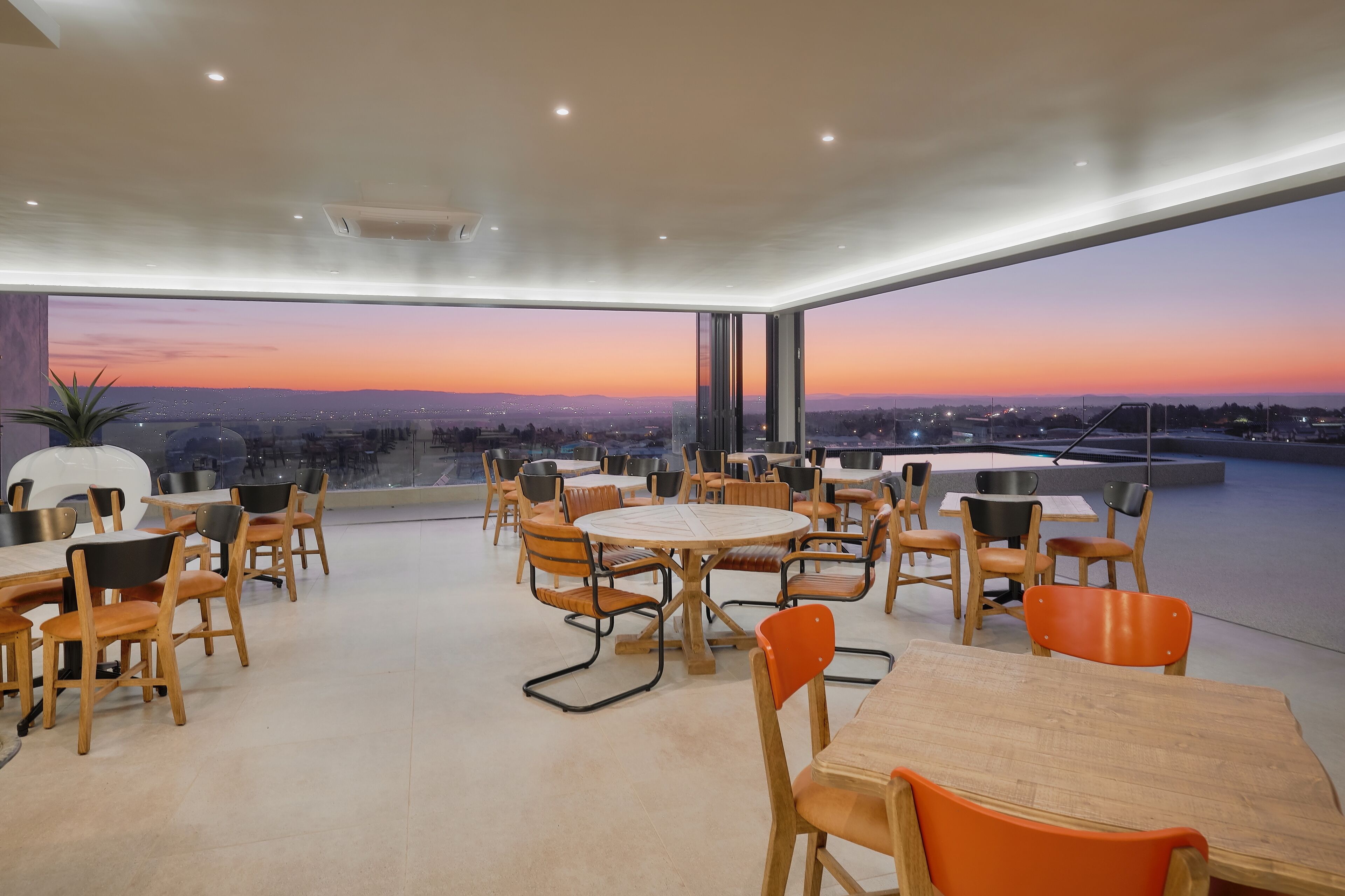 Ein moderner Essbereich mit runden Tischen und Stühlen, große Fenster mit Blick auf die Stadt bei Sonnenuntergang.