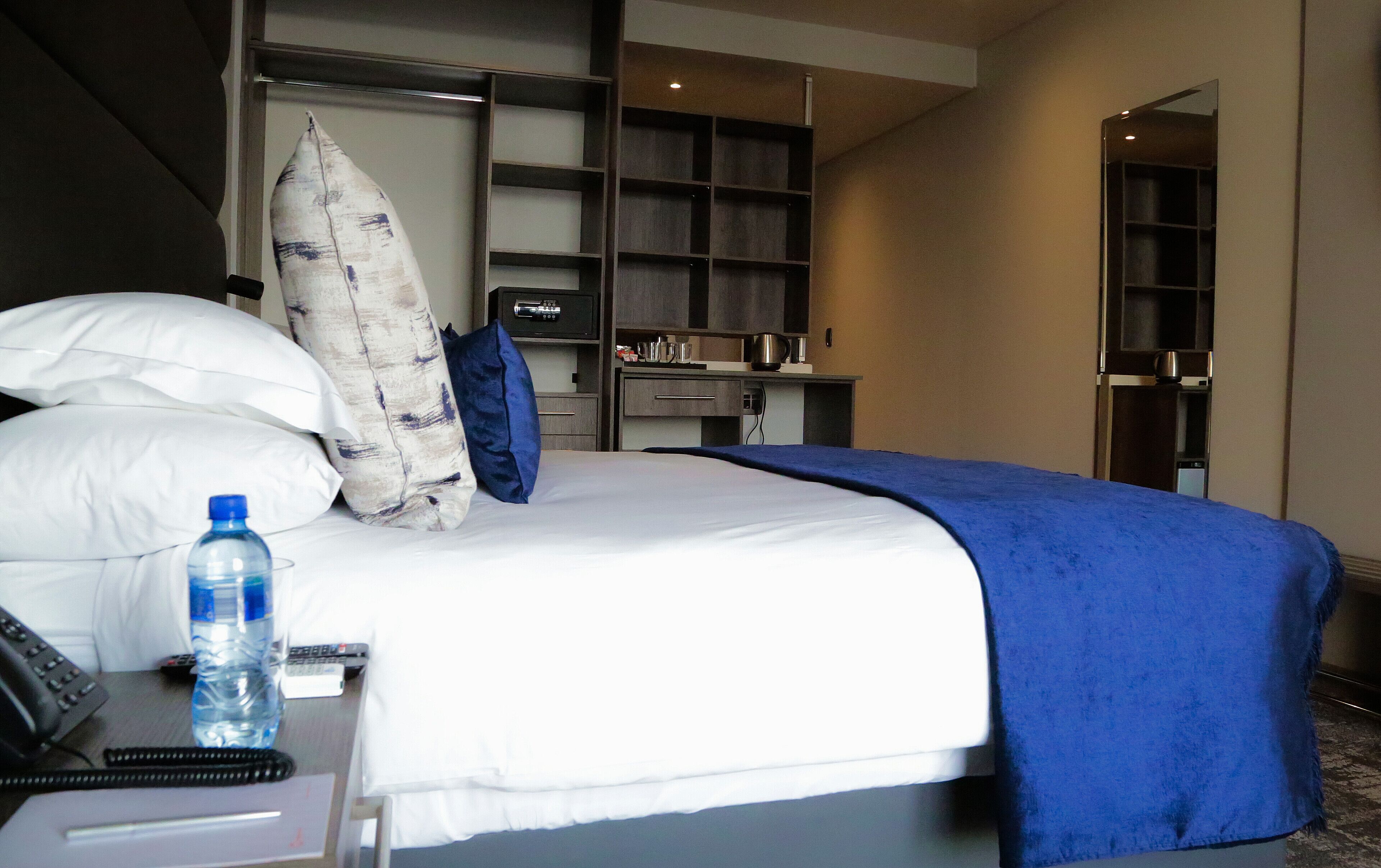 Une chambre d’hôtel comprenant un lit, un bureau avec un téléphone et une bouteille d’eau, un minibar et une salle de bain avec un miroir et un porte-serviettes.
