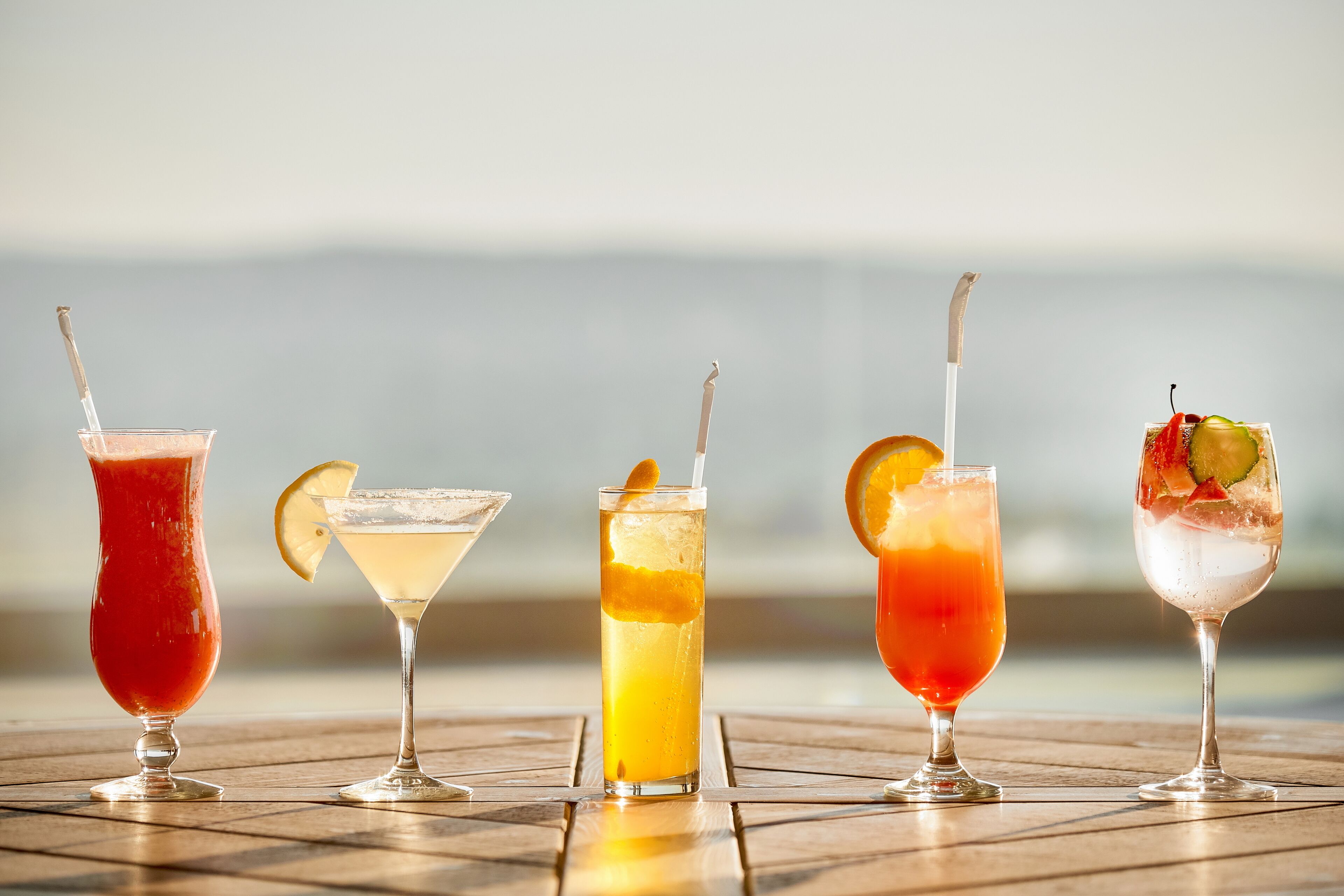Five cocktails on a wooden table with a blurred outdoor background.