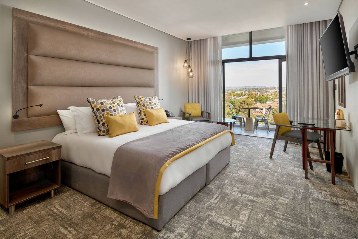 A modern hotel room with a large bed, a desk, and a chair. There is a TV mounted on the wall and a window with a view of the city.