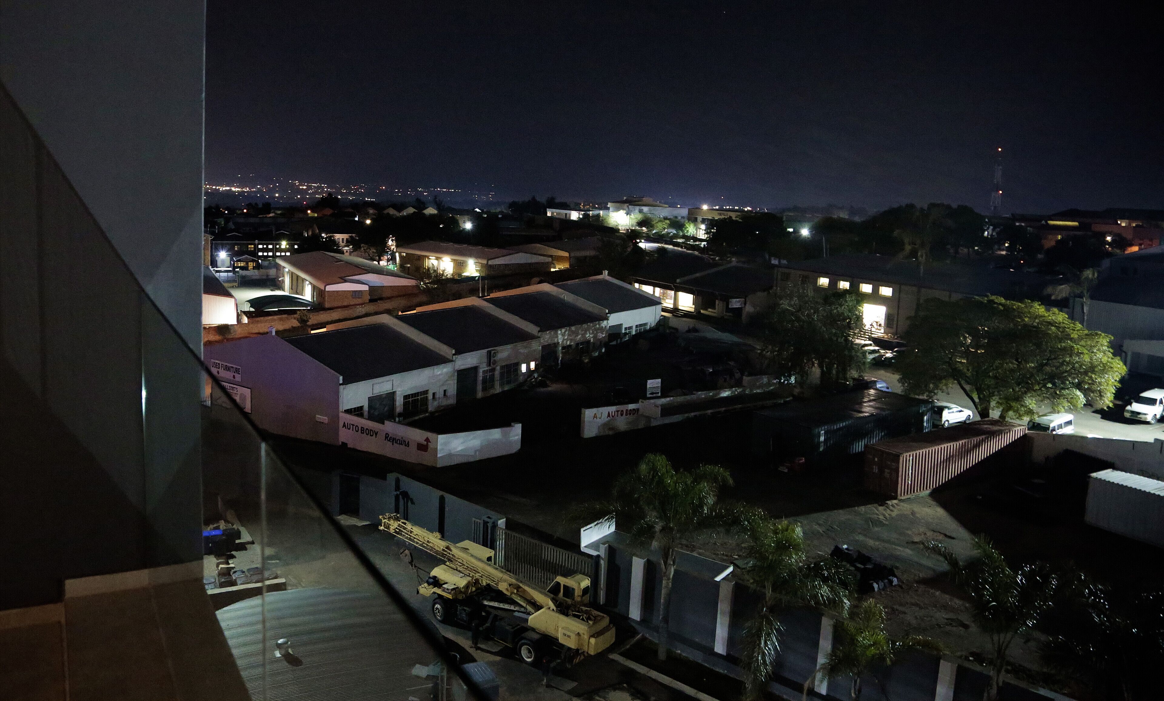 Vue nocturne d’une zone commerciale avec des immeubles, une grue et une enseigne indiquant « AJ AUTO BODY Repairs ».