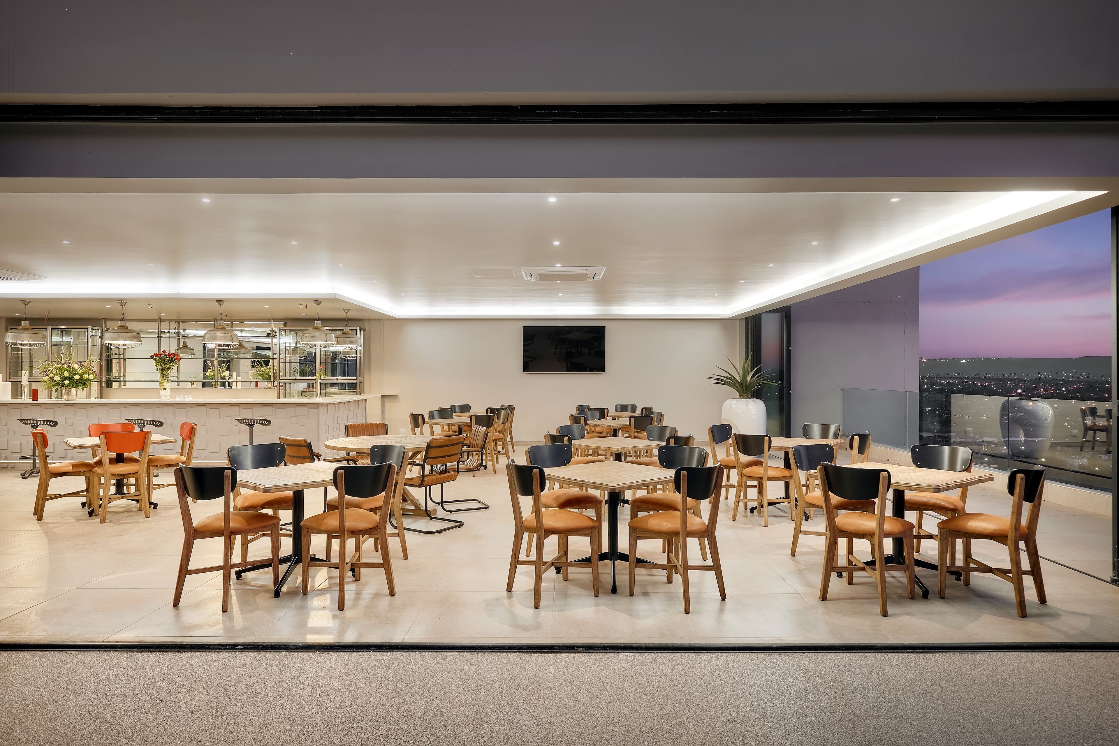 Un comedor moderno con varias mesas y sillas, una barra y un amplio ventanal con vista a la ciudad.