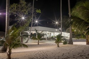 Un hotel frente a la playa con arena, palmeras y luces colgantes.