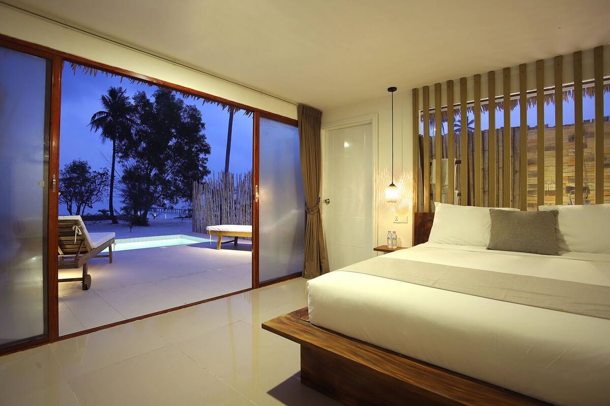 A modern bedroom with a large bed, a view of palm trees, and a sliding glass door leading to an outdoor area with a bench.