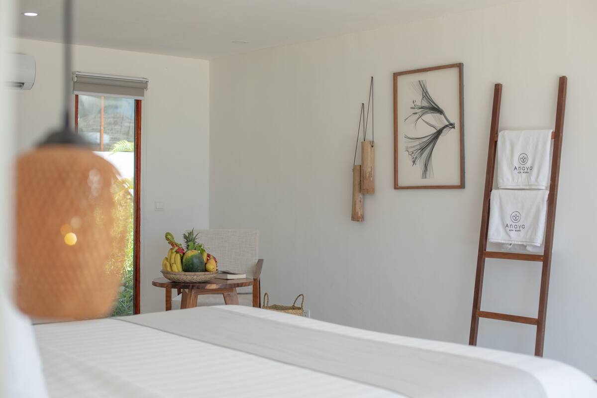 A bedroom with a bed, a wooden ladder, a table with fruits, and a wall with a framed artwork.