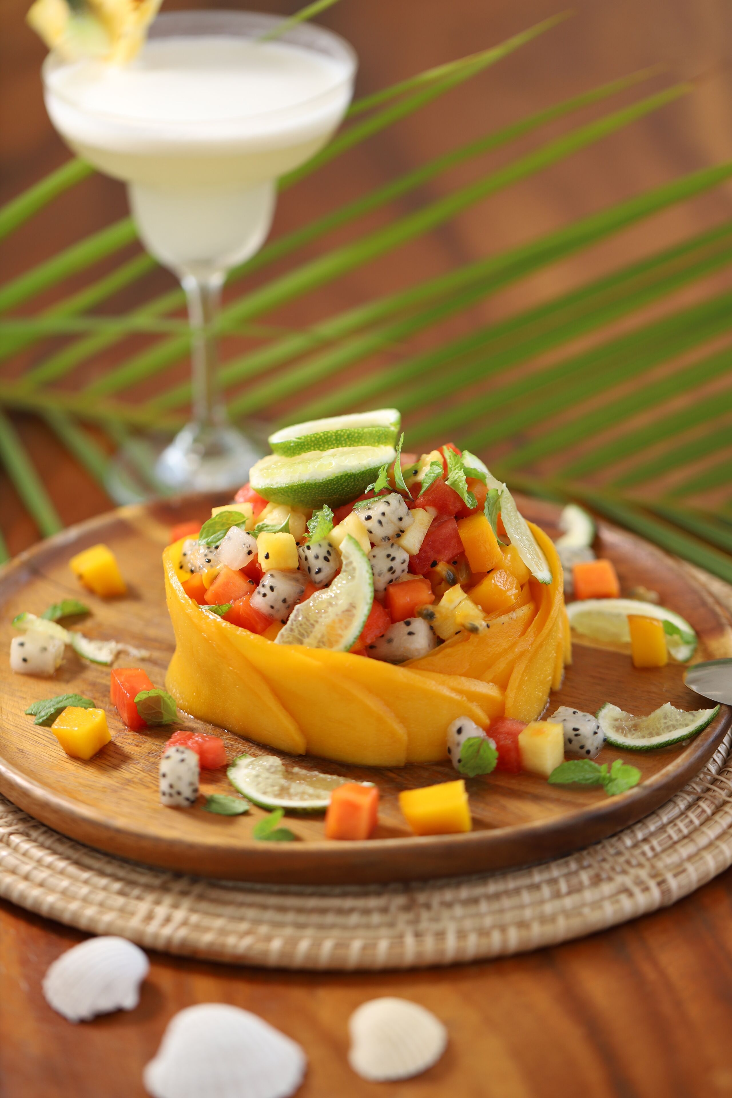 A fruit salad in a hollowed-out mango bowl on a wooden board, with a cocktail in the background.