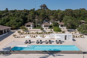 Un espace aménagé au bord de la piscine, avec des chaises longues et des parasols, entouré d’une plage de sable et bordé d’arbres et d’immeubles.