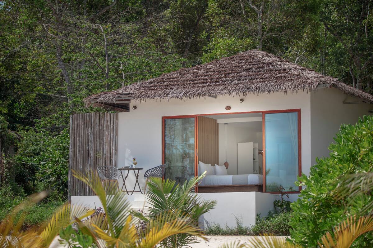 A small, single-story building with a thatched roof, large sliding glass doors, and a balcony with a table and chairs.