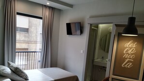 Un dormitorio con cama, televisor montado en la pared, baño visible a través de una puerta abierta y una lámpara colgante.