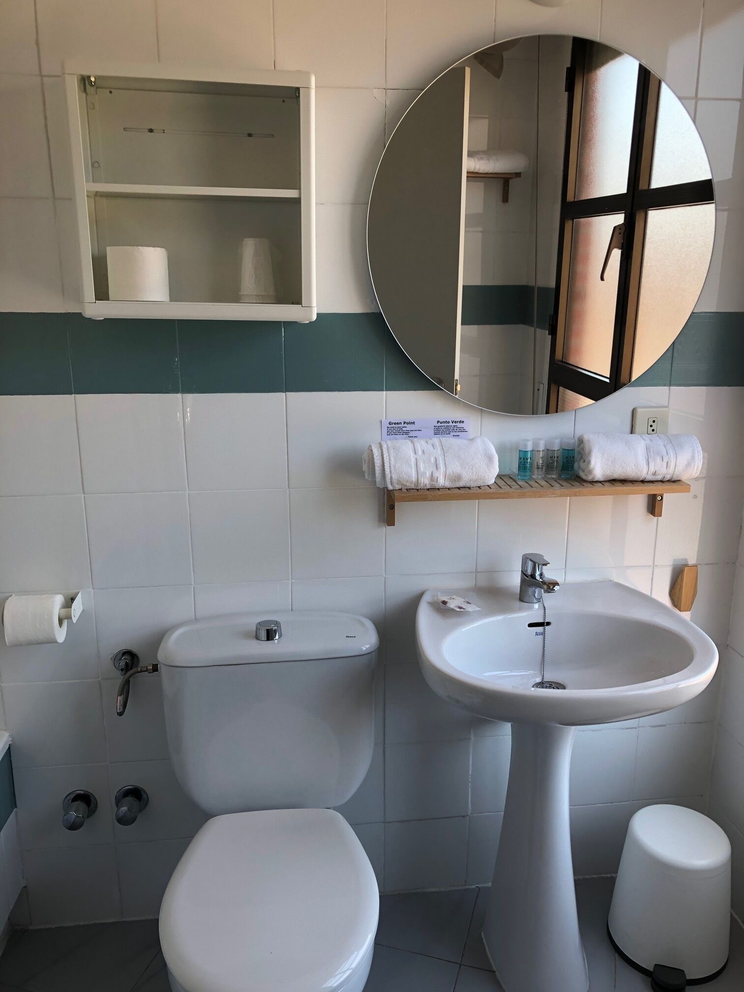 Baño con inodoro, lavabo y espejo. Hay un estante con artículos de aseo y una toalla.