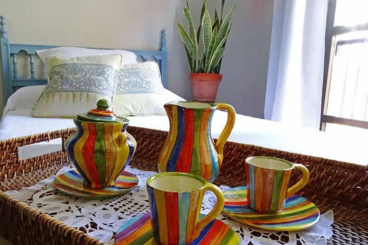 Een bed met een blauw hoofdbord en witte beddengoed, een potplant en een rieten dienblad met een theepot, karaf en kopjes.