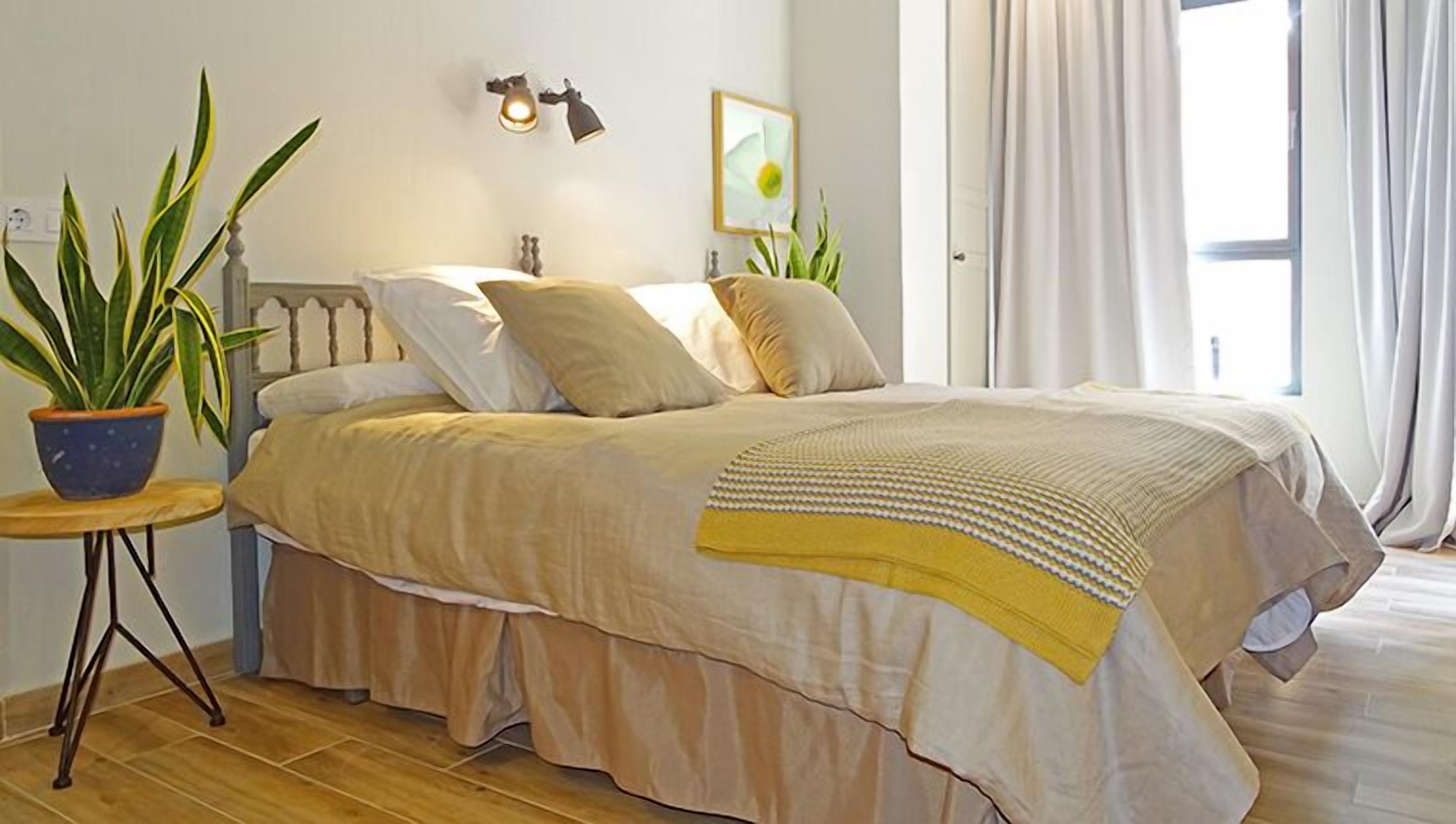Un dormitorio ordenado con una cama, una mesita de noche con una planta en maceta y una lámpara de pared.