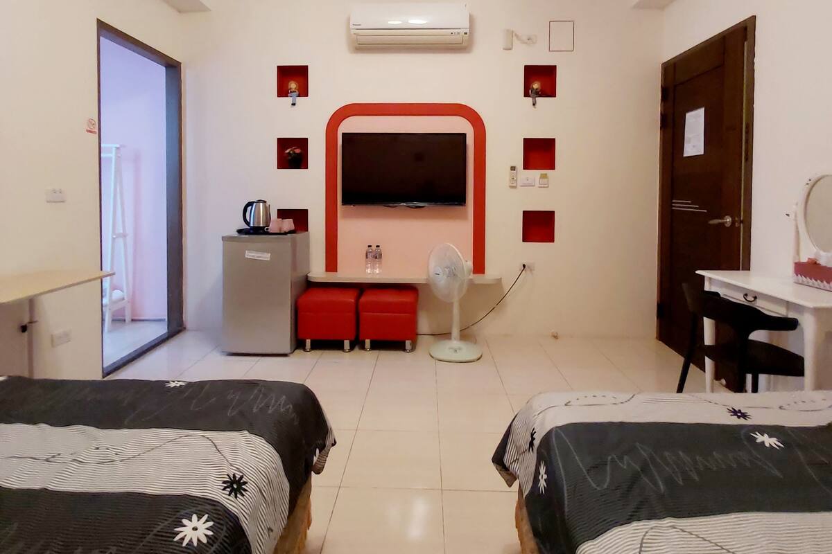 A hotel room with two beds, a desk, a chair, a small kitchenette, and a TV.