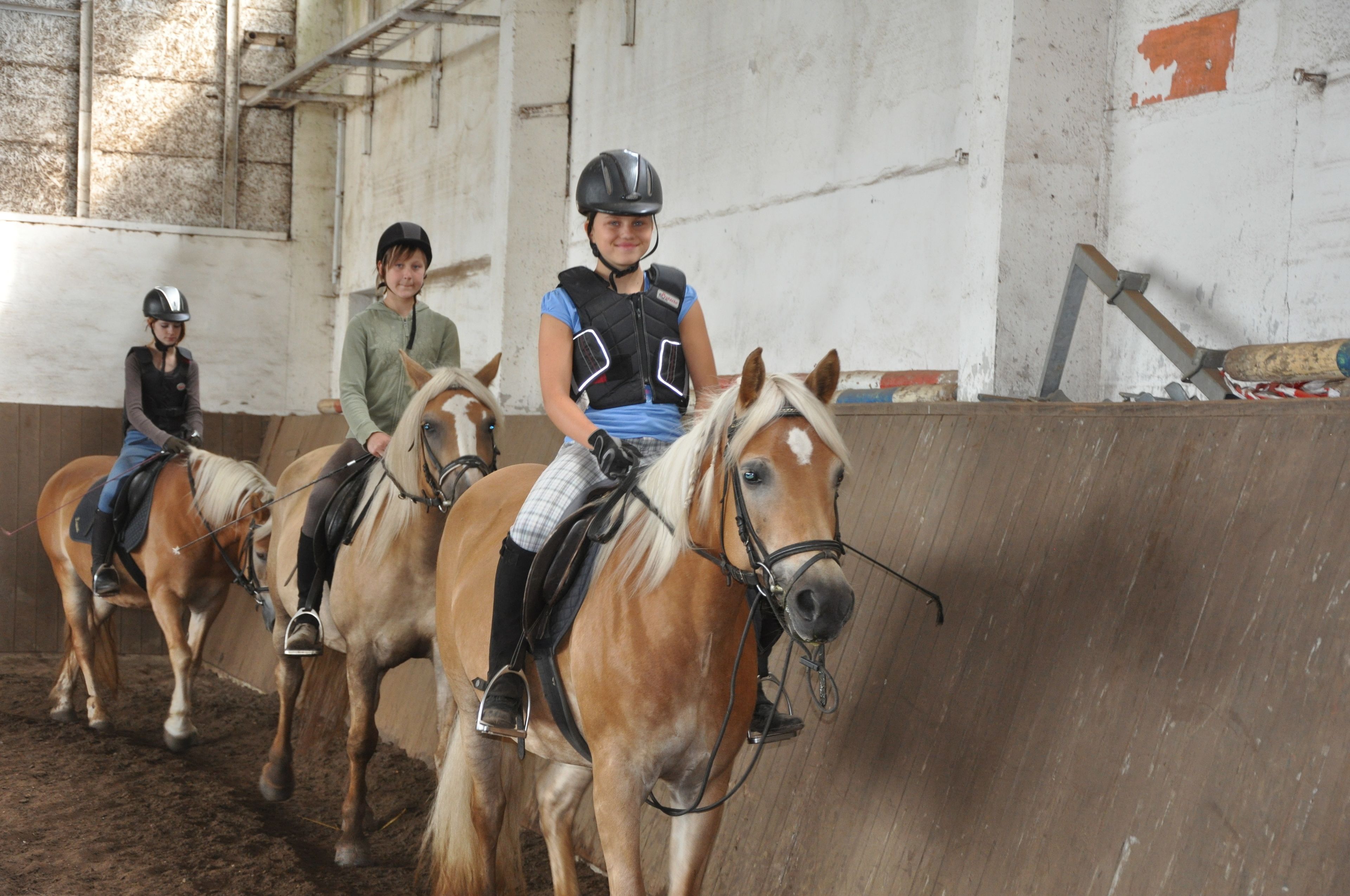 Three people riding horses in an indoor arena with a dirt floor and a concrete wall.