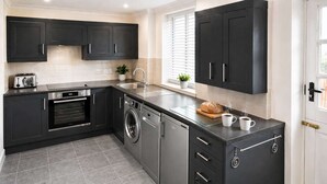 A modern kitchen with dark cabinets, a built-in oven, a washing machine, and a countertop with a toaster and a loaf of bread.