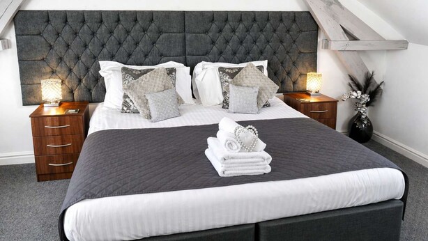 A neatly made bed with a tufted headboard, white and patterned pillows, and a stack of folded towels.