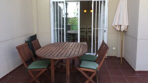 Un patio amb una taula de fusta rodona i quatre cadires amb coixins verds, una porta corredissa de vidre i un parasol blanc.