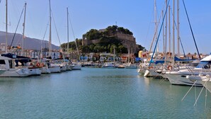 Un port esportiu amb diverses embarcacions amarrades, una imponent construcció semblant a un castell sobre un turó, i un cel blau i clar.