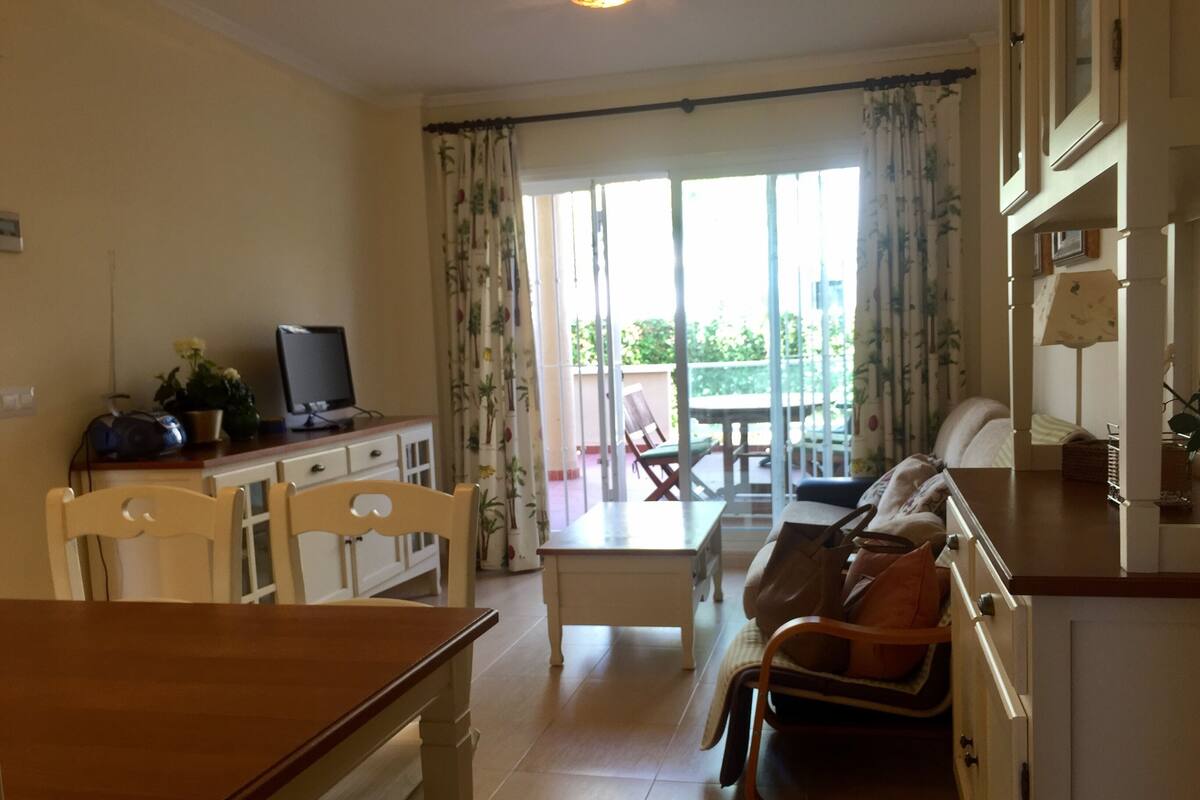 A living room with a wooden table, a sofa, and a TV. There is a balcony with a table and chairs.