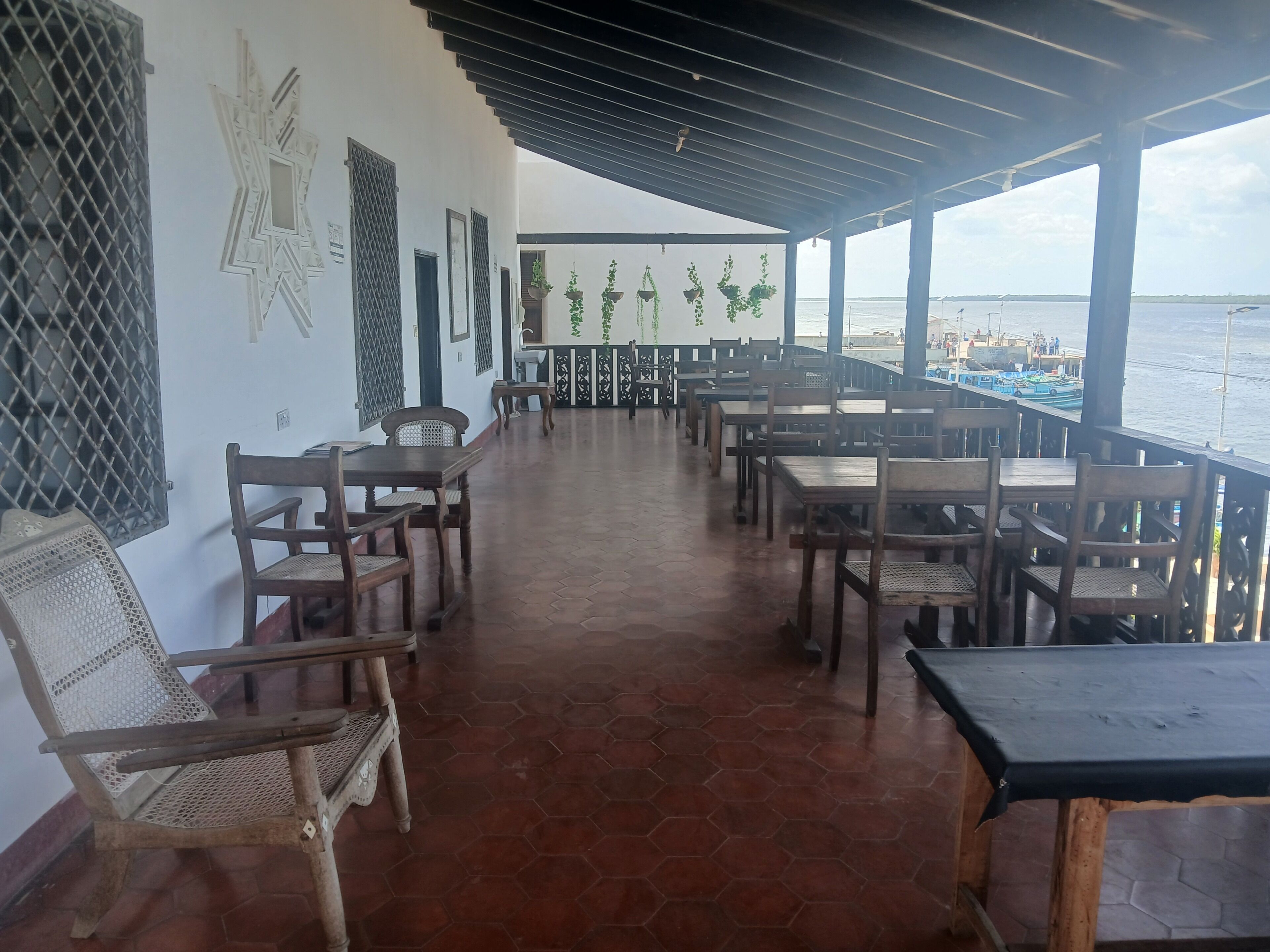 A covered outdoor seating area with hexagonal tiled flooring, wooden tables and chairs, and a view of the sea.