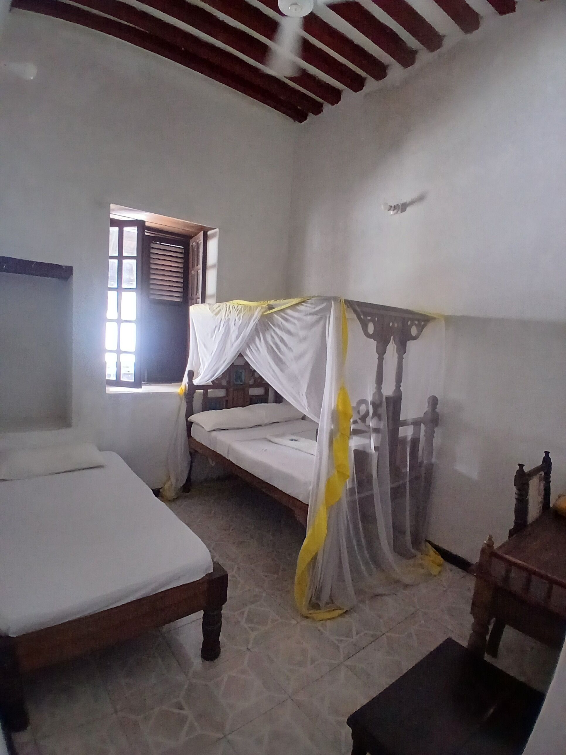 Une chambre avec deux lits, dont un à baldaquin, une fenêtre avec des stores et un ventilateur de plafond.