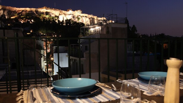 A dining table set with blue plates, glasses, and a white pitcher, overlooking a historic building at night. - Plaka 2-storey Penthouse with Acropolis views. PL6 (Athens)