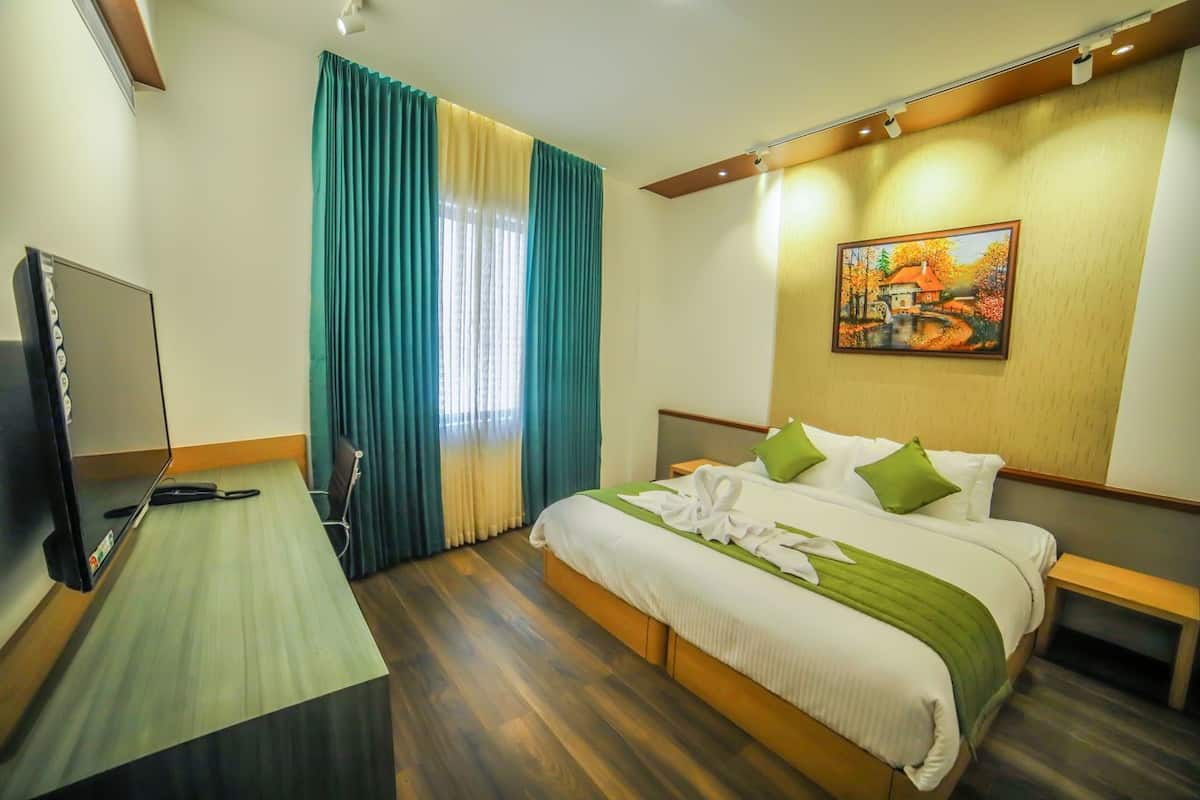 A hotel room with a bed, a desk with a television, a painting on the wall, and a window with curtains.