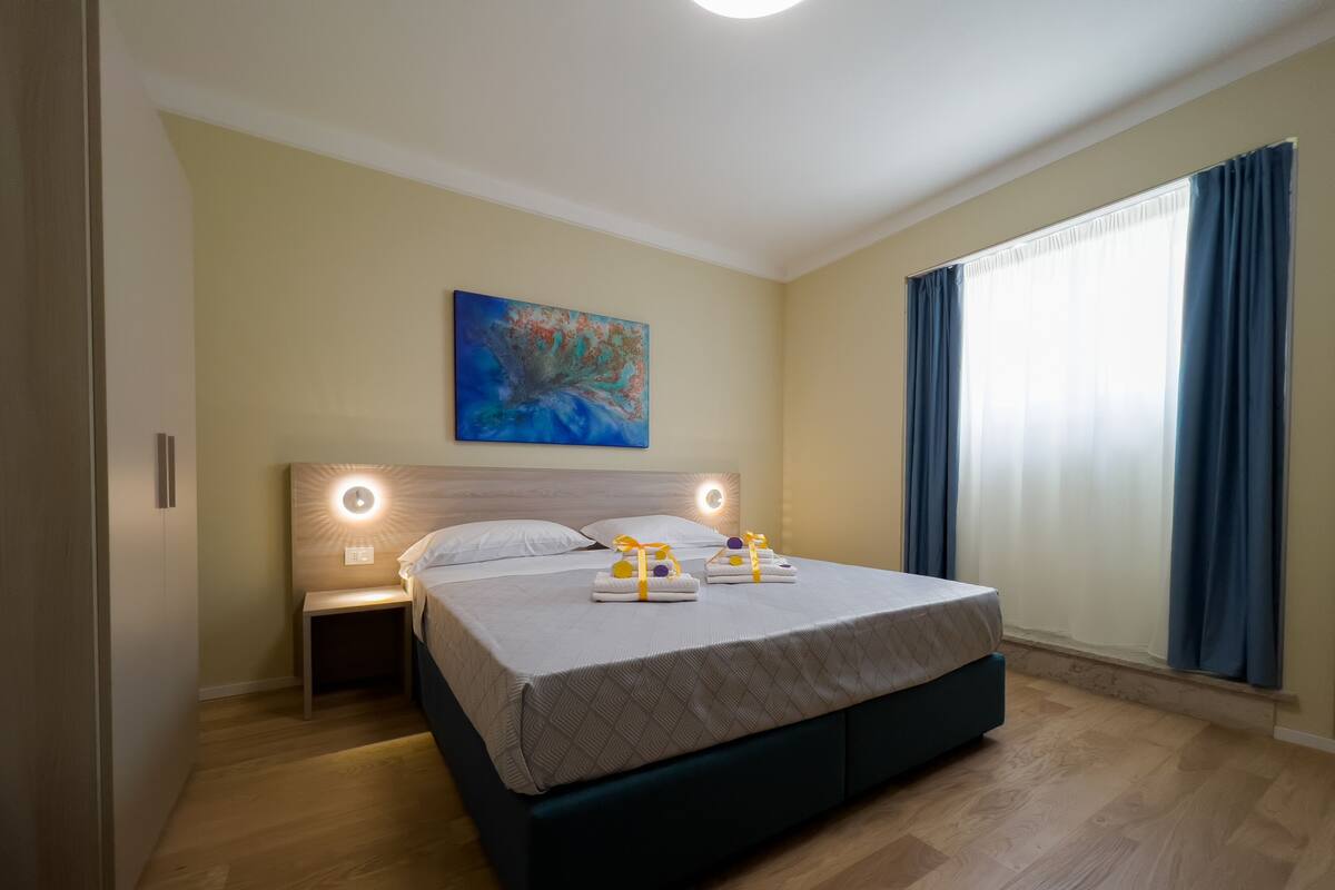 A modern hotel room with a large bed, bedside lamps, a painting on the wall, and a window with curtains.