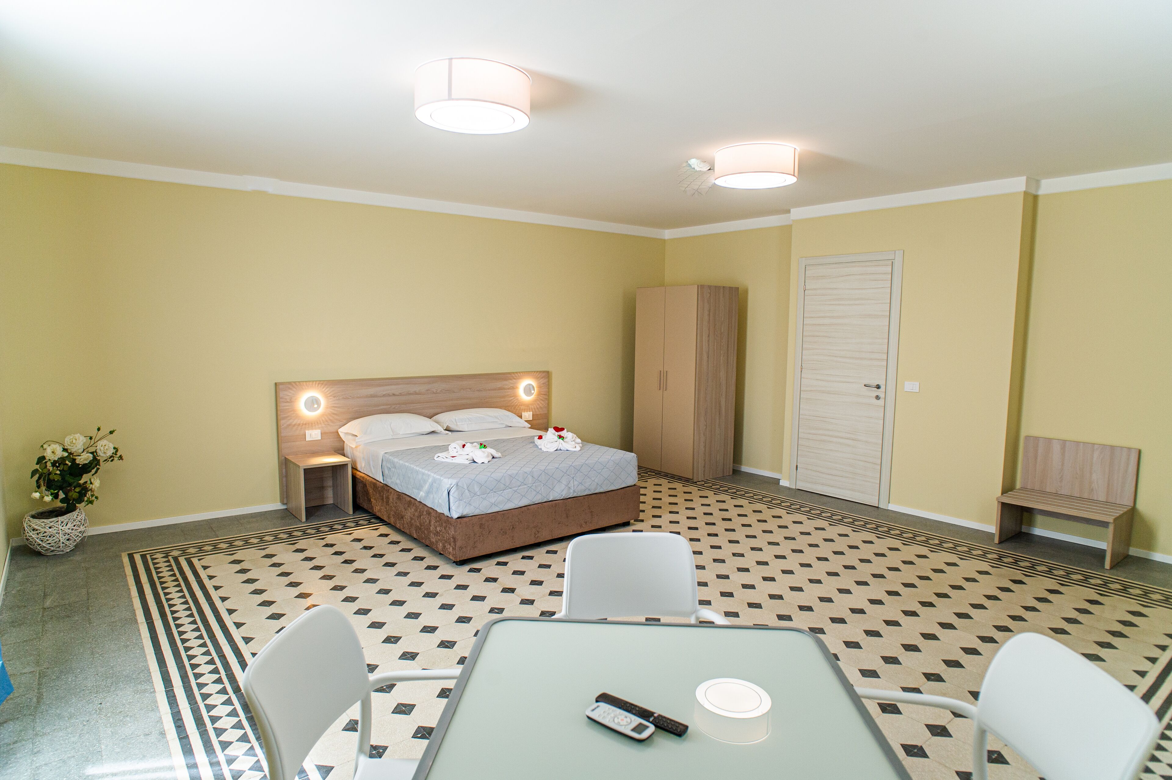 A hotel room with a bed, bedside table, lamp, chair, table, and a small bench.