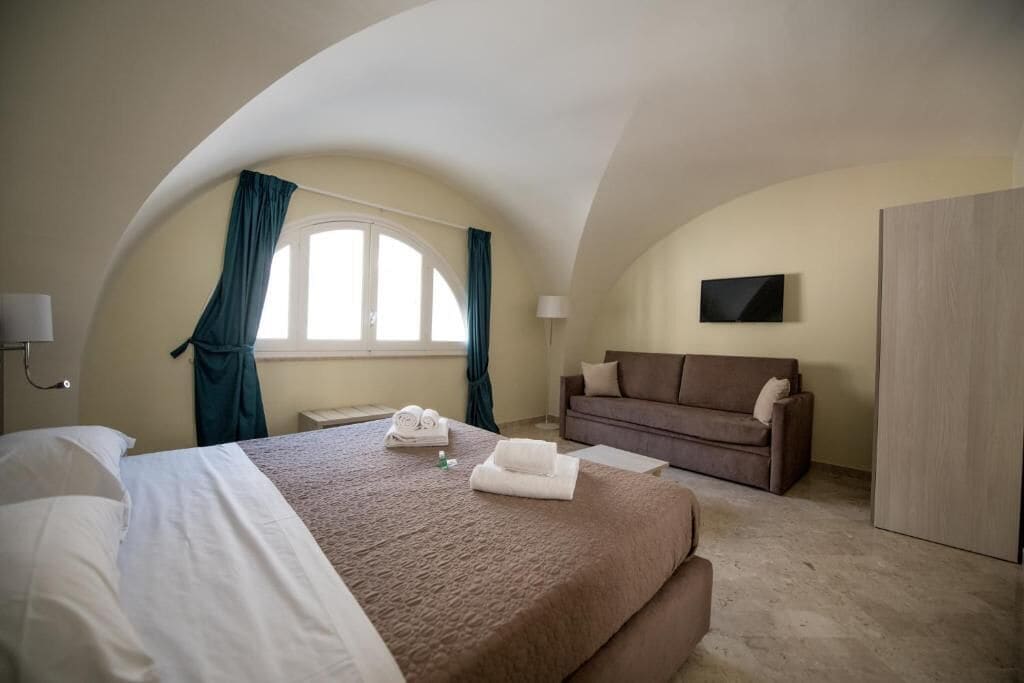 Habitación de hotel con una cama grande, un sofá, televisión montada en la pared y una ventana con cortinas.