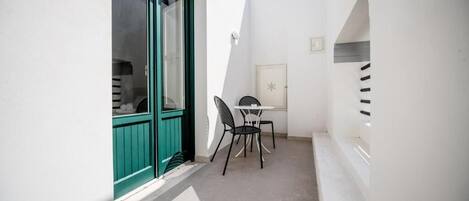 Un estrecho pasillo exterior con una pared blanca, una puerta verde y una pequeña mesa con dos sillas.