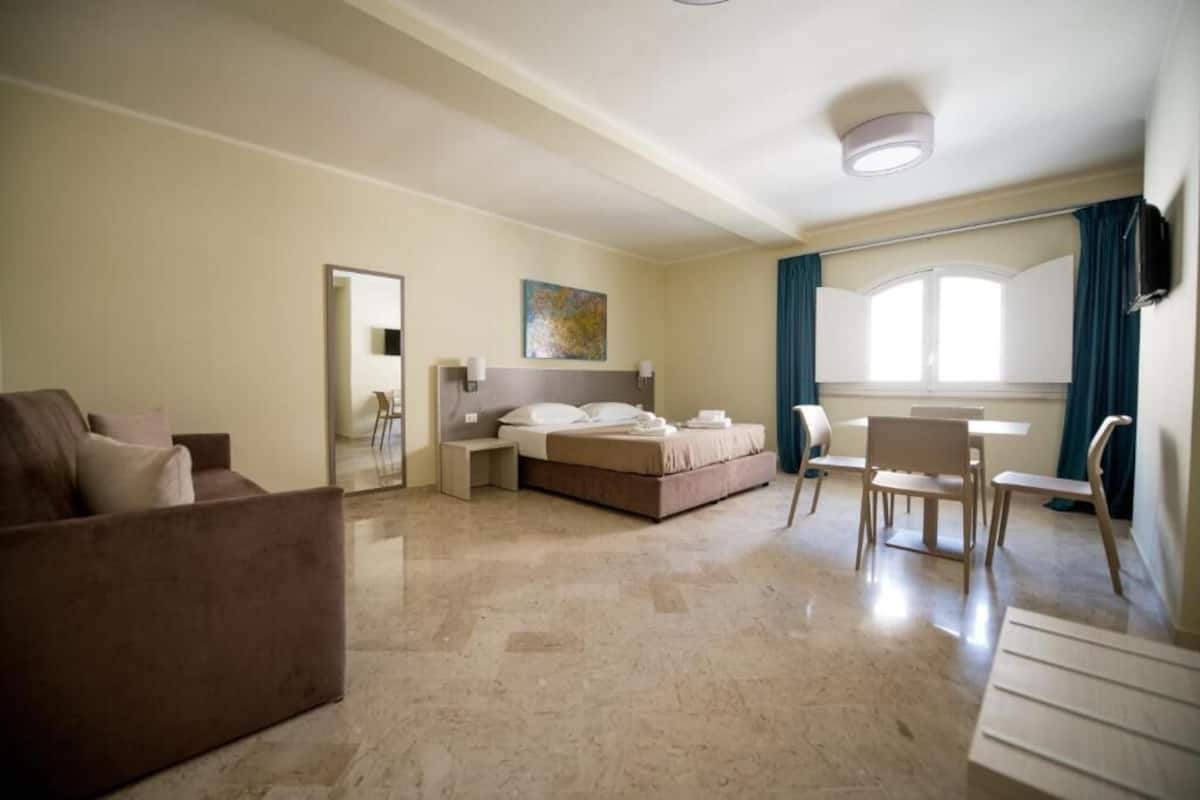 A hotel room with a bed, a sitting area with a sofa and chairs, a dining table, and a large window with curtains.