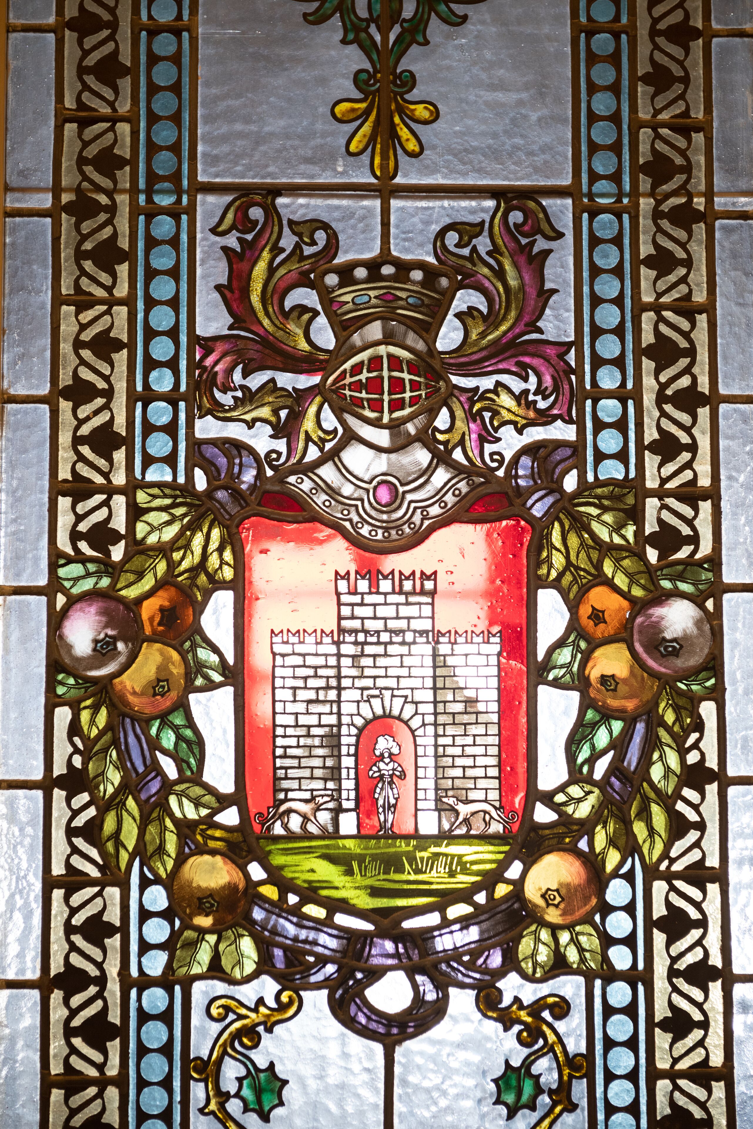 Une fenêtre en vitrail ornée d’un écu central représentant un château et une figure, le tout étant entouré de motifs décoratifs de feuillage et de fruits.
