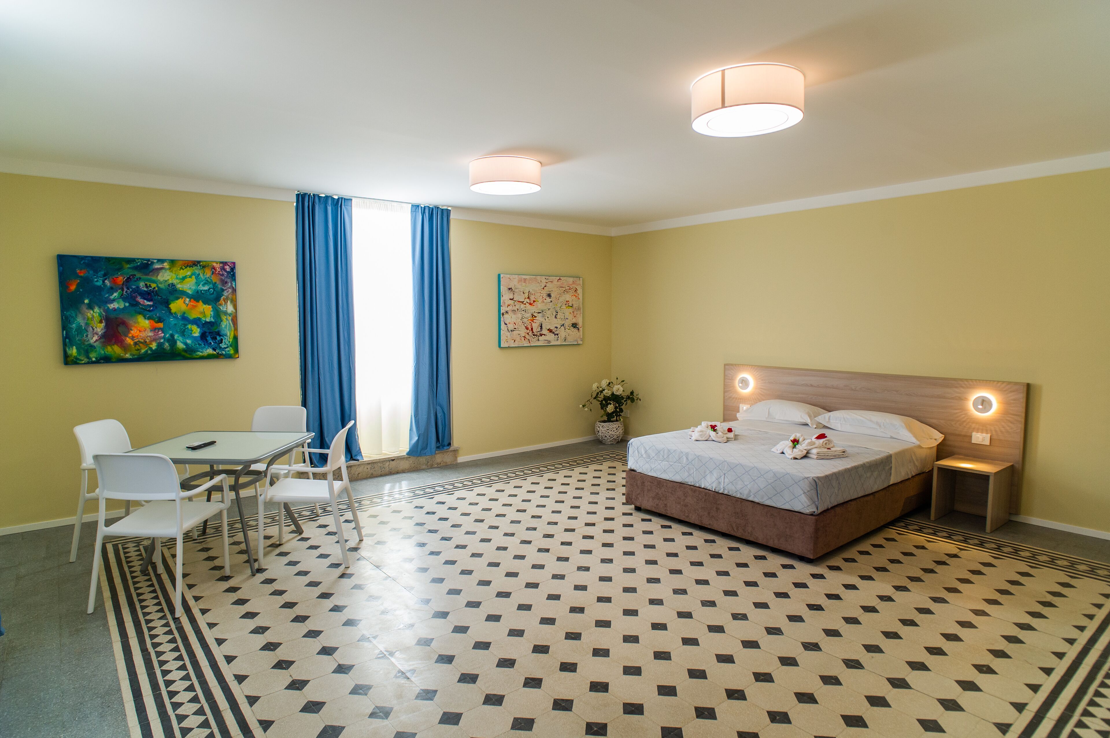 Une chambre d’hôtel avec un lit, un bureau avec des chaises, une fenêtre avec des rideaux et deux tableaux accrochés aux murs.