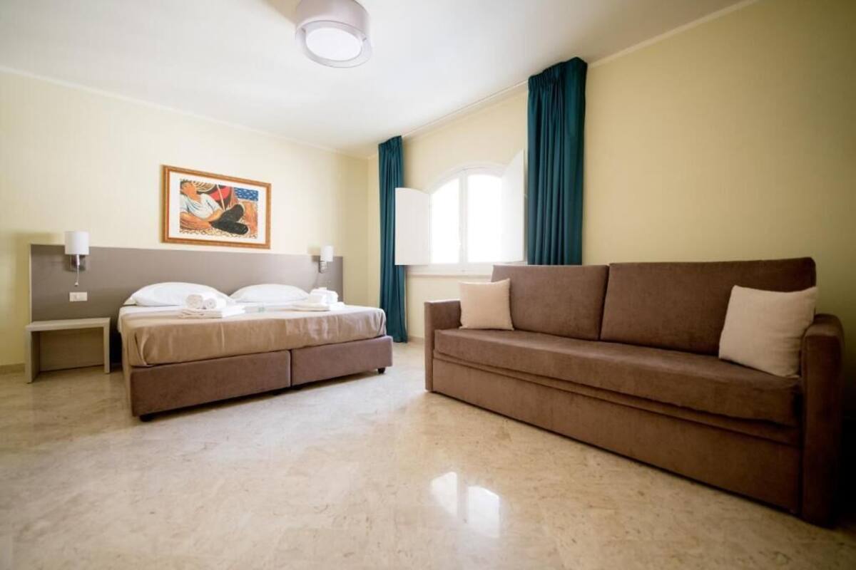 A hotel room with a bed, a sofa, a painting, and a window with curtains.