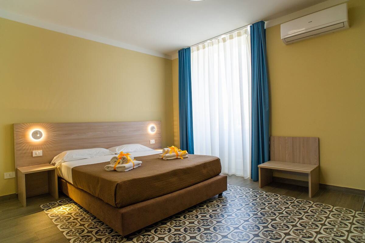 A hotel room with a large bed, bedside lamps, a bench, and a patterned rug.